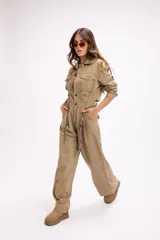 Mono de estilo utilitario color beige, con cierre frontal de botones, bolsillos en el pecho y cintura entallada con detalle de cadenas decorativas. Presenta mangas largas y piernas de corte recto y holgado.