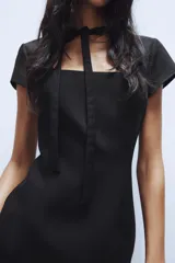 Vestido corto negro de cuello redondo con lazada, escote cuadrado y manga corta, con cierre en la espalda con cremallera oculta.