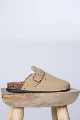 Zueco de gamuza sintética color beige, con detalle de tachas metálicas doradas y hebilla lateral ajustable. Presenta una base de corcho y suela de goma dentada.