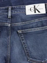 Jeans de hombre de mezclilla azul oscuro con efecto lavado y arrugas marcadas en la parte delantera. Presentan un corte slim, tiro alto, cierre de cremallera y botón, y diseño clásico de cinco bolsillos. Incluyen un parche con logo en la cintura trasera.