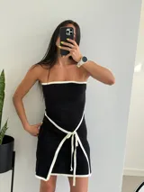 Vestido corto negro sin mangas, con escote recto con borde blanco y lazo blanco decorativo en la cintura.