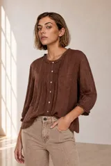 Blusa de manga larga color marrón, con cuello redondo y detalles fruncidos con volados pequeños en el cuello y los puños. Presenta cierre frontal con pequeños botones blancos y delicados pliegues verticales en el pecho.
