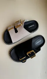 Sandalias tipo chinela marca Vdamiani, confeccionadas en nobuck con una tira ancha ajustable mediante hebilla metálica dorada. Presentan una base negra con detalle de costura perimetral en contraste y plantilla ergonómica.