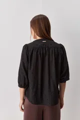 Camisa negra de textura liviana con detalle de lunares en relieve, cuello mao y cierre frontal con botones. Presenta mangas 3/4 con puños elastizados y frunces en los hombros.