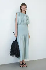 Vestido midi de tejido texturizado color verde menta, con escote redondo fruncido, espalda semiabierta con breteles finos y capa decorativa sobre los hombros. Cuenta con sección de punto smock en la cadera y falda con leve vuelo.