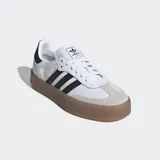 Zapatillas Adidas Sambae blancas con las tres tiras laterales en negro, refuerzo de gamuza gris en la puntera, contrafuerte trasero negro y suela de goma color caramelo.