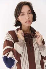 Buzo de tejido de punto con diseño de rayas verticales en tonos bordó y rojo. Presenta cuello alto con cierre frontal, mangas largas con coderas en contraste y detalles de costuras visibles.