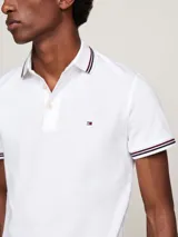 Chomba blanca de algodón elástico, corte slim, con cuello y puños acanalados con ribetes en azul marino y rojo. Tiene cartera de tres botones y logo de Tommy Hilfiger bordado en el pecho.