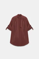 Camisa bordó de cuello alto ajustable con doble hebilla y cierre frontal con botones ocultos por solapa. Tiene mangas cortas con trabillas y diseño holgado.