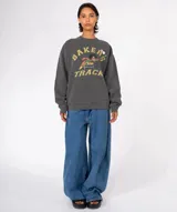 Buzo de algodón y poliéster color gris oscuro lavado, con cuello redondo y puños y ruedo acanalados. Presenta un estampado frontal con el texto "BAKERS" y "TRACK" en amarillo y verde, y un dibujo de un pato saltando una valla. Tiene dos pines decorativos prendidos en el área del pecho.