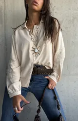Camisa de corte holgado en color beige, con cuello clásico, cierre frontal de botones y mangas largas que presentan un delicado bordado tonal en los antebrazos.