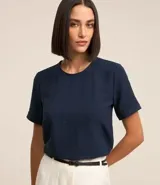 Blusa de tejido plano color azul marino, con cuello redondo, mangas cortas y detalle de pespuntes en cuello y mangas. Presenta un cierre trasero tipo gota con botón y pequeñas aberturas laterales.