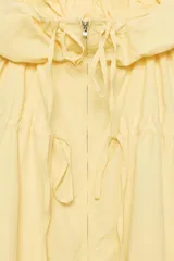 Chaqueta de nylon color amarillo pastel, con cuello subido y capucha. Presenta cierre frontal con cremallera, mangas largas con puños elásticos y detalle de lazadas ajustables en el frente. El bajo cuenta con cordón elástico y stoppers para un ajuste personalizado.