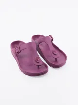 Sandalias tipo ojota de goma, con diseño de dedo y tira ajustable con hebilla lateral. Presentan una plantilla anatómica y un pequeño aplique circular decorativo en la parte frontal.