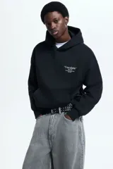 Canguro de corte oversized en mezcla de algodón, color negro, con capucha de doble capa, hombros caídos y bolsillo frontal tipo canguro. Presenta un estampado tipográfico pequeño en el pecho y puños y bajo acanalados.