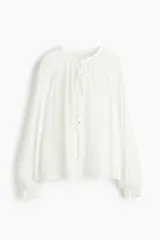 Blusa de crepé de gasa color blanco, con cuello mao, abertura frontal con tiras para anudar y detalle de volados verticales. Presenta mangas largas tipo globo de corte raglán con puños elásticos y terminación en volados.