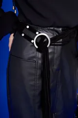 Cinturón negro de cuero con hebilla metálica plateada en forma de aro. El cinturón se anuda y tiene una tira de gamuza negra con flecos colgantes.