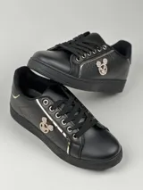 Zapatillas deportivas de cuero sintético en color blanco, beige y negro, con cordones y aplique decorativo dorado con forma de moño.