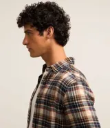 Camisa de flanela con estampado a cuadros en tonos beige, azul y rojo. Presenta cuello clásico, cierre frontal con botones y mangas largas con puños abotonados.