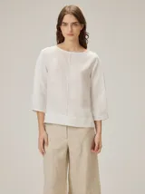 Blusa de algodón color blanco crudo, de corte recto, con cuello redondo y mangas tres cuartos abullonadas. Presenta una costura vertical central en el frente.