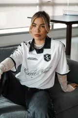 Camiseta de fútbol estilo boxy color blanco, con cuello polo y ribetes negros en las mangas. Presenta detalles estampados y bordados exclusivos en el frente y hombros.