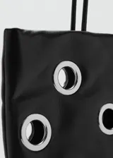 Bolso tipo shopper de color negro con diseño de ojales metálicos plateados. Cuenta con doble asa de hombro y cierre de botón magnético.