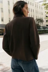 Blazer de corte estructurado en color marrón chocolate, con cuello solapa, hombros marcados y cierre frontal cruzado con botón oculto. Presenta un bolsillo superior tipo ojal y un diseño de bajo redondeado.
