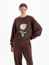 Buzo oversize color marrón de cuello redondo y manga larga, con estampa frontal de una rosa blanca y logo bordado en la espalda.