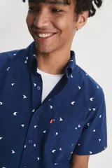 Camisa de manga corta color azul con estampado de pájaros blancos, cuello clásico con botones y bolsillo frontal con etiqueta roja.