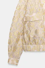 Chaqueta tipo bomber de cuello redondo y manga larga, confeccionada en tejido jacquard con diseño floral en tonos dorados y crema. Presenta cierre frontal con cremallera, bolsillos delanteros de plastrón con solapa y puños y bajo elásticos.