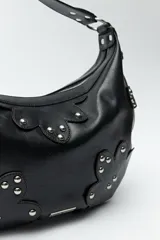 Cartera marrón de cuero sintético con forma de media luna, asa corta y apliques de tachas metálicas plateadas.