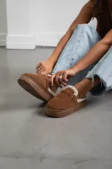 Zueco de gamuza color marrón, con interior forrado en corderito sintético, suela de plataforma plana y detalle de tira con hebilla metálica en el empeine.