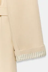 Abrigo largo de lana en color beige, con cuello de solapa, cierre cruzado con botones y cinturón ajustable del mismo tejido. Presenta mangas largas con detalle en los puños y bolsillos de vivo en el delantero.
