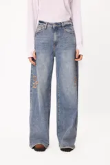 Jean de corte wide leg en denim azul claro con efecto lavado. Presenta bordados del logo de la marca en color ocre en ambos laterales a la altura de las rodillas y ruedo desflecado.