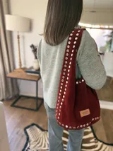 Bolso tipo tote de gamuza color bordó, con tachas plateadas en el contorno y en la correa. Tiene un parche de cuero con el logo de la marca en el frente.