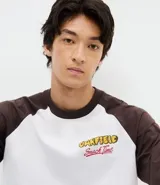Remera de algodón con diseño estilo baseball, cuerpo blanco y mangas cortas en color marrón oscuro. Presenta un estampado gráfico colorido en la espalda con el personaje Garfield rodeado de comida y elementos decorativos.