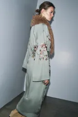 Kimono de largo midi en color verde grisáceo, con cuello de solapas amplias en pelo sintético marrón. Presenta mangas largas acampanadas y un cinturón ajustable del mismo tejido. El diseño está decorado con un delicado bordado floral en tonos tierra y rosados.