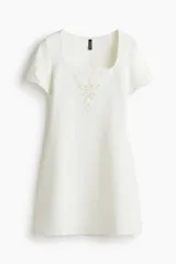 Vestido corto color marfil, confeccionado en punto interlock pesado, con escote cuadrado, mangas cortas y detalles de inserciones bordadas en el busto y las mangas.
