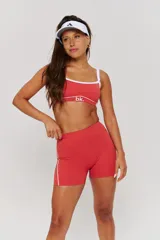Conjunto deportivo compuesto por un top y un short de tiro alto en color bordó con detalles de líneas blancas. El top presenta breteles anchos y el logo de la marca en el frente, mientras que el short tiene un diseño ajustado con costuras laterales visibles.