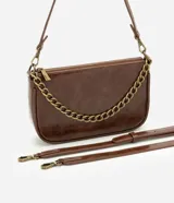 Cartera femenina modelo baguette Satinato confeccionada en PU marrón, de tamaño pequeño y estructura compacta con base levemente curvada. Cuenta con cierre superior, cadena frontal robusta en metal y asa transversal desmontable.