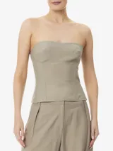 Top corset color beige, sin tirantes, confeccionado en tela ligera no elástica. Presenta escote recto, aberturas delanteras y traseras, largo corto, bajo asimétrico y cierre de cremallera en la espalda.