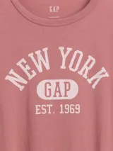 Remera de manga corta con cuello redondo, color rosado, con estampa frontal con texto New York Gap Est. 1969 en color blanco.