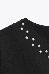 Top negro de manga corta con cuello redondo, adornado con perlas blancas aplicadas en el escote y en los puños de las mangas. Cierre posterior con botones de perla.