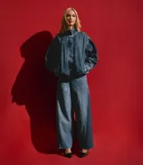 Pantalón wide leg de jean azul, con cintura media, trabillas para cinturón, cierre frontal tradicional y bolsillos. Presenta un diseño con recorte lateral en un tono de azul más oscuro que contrasta con el resto de la prenda.
