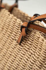 Bolso de mano tipo tote confeccionado en tejido texturizado efecto rafia en color beige, con asas dobles finas de cuero sintético marrón y detalles de nudos en los extremos.