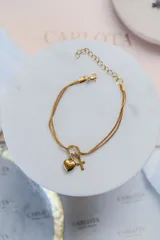 Pulsera de doble cadena fina en acero quirúrgico con baño de oro 18k. Presenta un nudo central del cual cuelgan dos dijes: un corazón liso y una cruz pequeña.