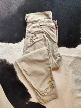 Bombacha de campo de lino color beige, con bordados florales en tono naranja y verde en el bolsillo delantero y en el botamanga.