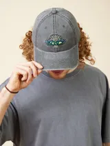 Gorro de gabardina gris oscuro con visera curva, ajustable en la parte trasera con hebilla metálica. Presenta bordado frontal con el logo de la marca y el año 1985.