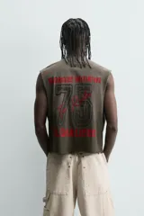 Camiseta tipo tank (sin mangas) de corte cropped fit, color gris oscuro con efecto gastado. Presenta cuello redondo y un pequeño parche rectangular con texto en la parte inferior delantera.