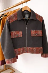 Campera de corte oversize confeccionada en lana merino, con diseño bicolor que combina paneles de color gris oscuro liso con detalles en tejido de punto con textura y estampado a cuadros en tonos rojizos y marrones. Presenta cierre frontal, cuello camisero, bolsillos frontales con solapa y puños con el mismo tejido estampado.